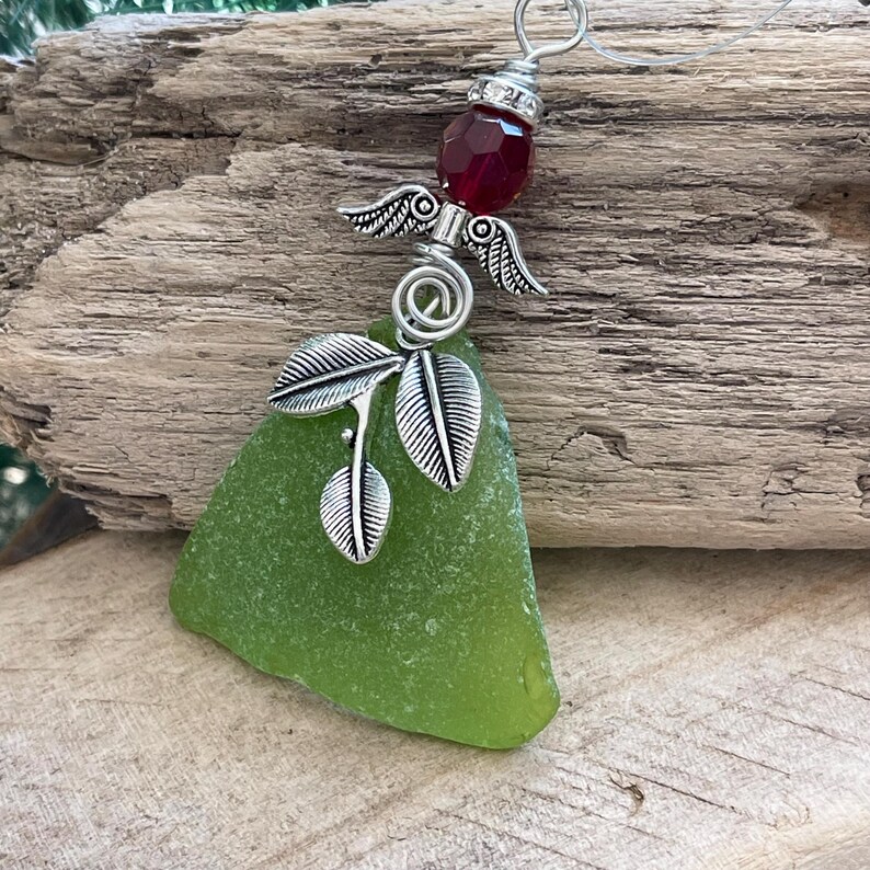 Pode incluir: Pingente artesanal com um peda&ccedil;o de vidro do mar em um tom verde vibrante. O pingente &eacute; adornado com um pingente de asas de anjo prateado, uma pedra preciosa vermelha e delicados detalhes de folhas. O design &eacute; suspenso por um fio de prata.