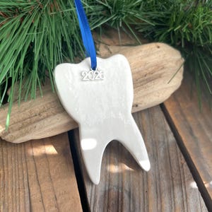 May include: A white ceramic tooth-shaped ornament with a glossy finish, suspended by a bright blue ribbon. A small silver-toned metal charm featuring the year 2026 is attached to the ribbon loop at the top of the tooth-shaped ornament.
