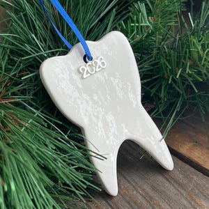May include: A white ceramic tooth-shaped ornament with a glossy, mottled finish. A silver-toned metal charm with the year 2026 is attached to the top, suspended by a thin blue ribbon. The ornament has two distinct roots at the base.