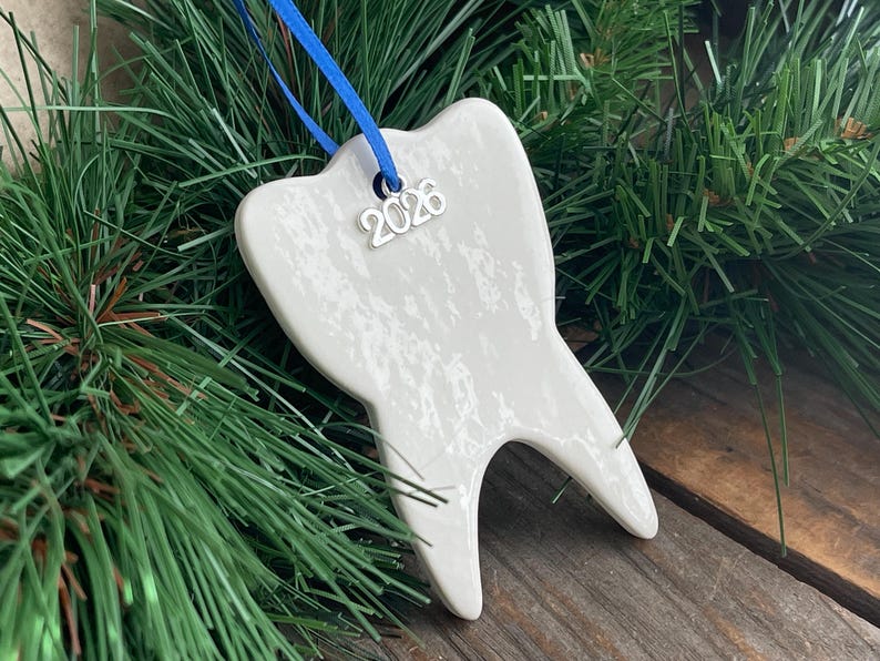 May include: A white ceramic tooth-shaped ornament with a glossy, mottled finish. A silver-toned metal charm with the year 2026 is attached to the top, suspended by a thin blue ribbon. The ornament has two distinct roots at the base.