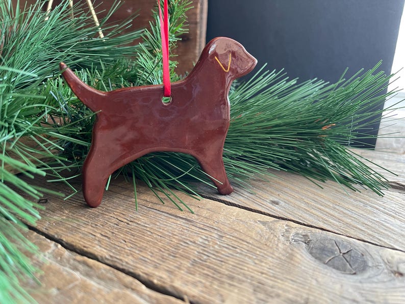 Pu&ograve; includere: Un ornamento in ceramica marrone scuro lucido a forma di cane labrador. La figura presenta una superficie liscia e riflettente con un piccolo contorno giallo dell'orecchio. Un sottile nastro rosso &egrave; infilato in un foro nella parte superiore.