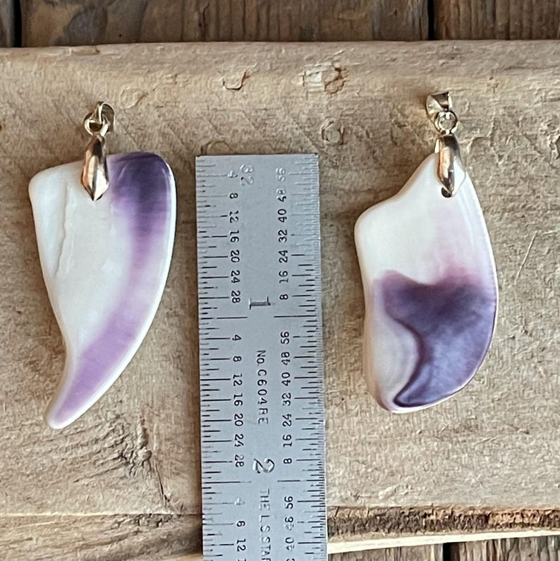 May include: Two pendant necklaces with a white and purple design. The pendants have a gold-colored bail at the top. A ruler is placed between the pendants to measure their length.