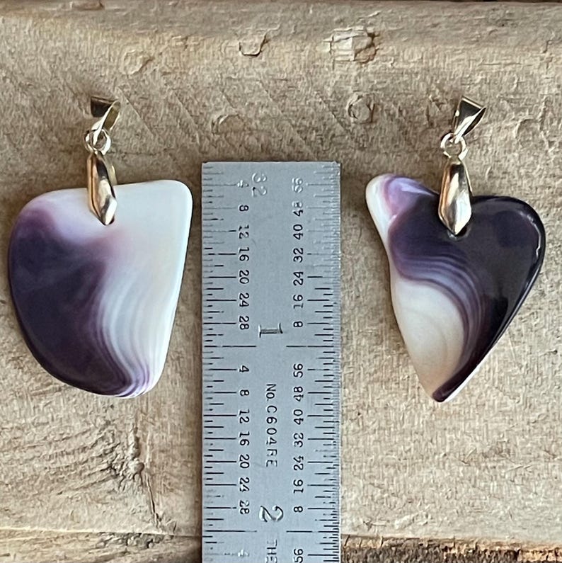 May include: Two pendants with a purple and white swirl pattern, each with a silver-colored bail. The pendants are approximately 1.5 inches long. The background is a light brown wooden surface.