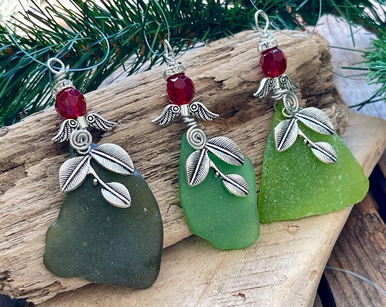 Pode incluir: Tr&ecirc;s enfeites de vidro do mar com asas de anjo prateadas e detalhes de folhas. Cada enfeite tem um tom diferente de vidro do mar verde, com uma conta vermelha no topo. Os enfeites s&atilde;o exibidos em um peda&ccedil;o de madeira flutuante.