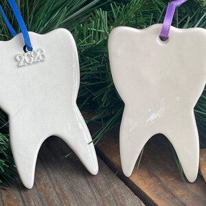 May include: Two white ceramic tooth-shaped ornaments. One ornament features a blue ribbon and a silver charm with the text 2026. The other ornament features a purple ribbon. Both items have a smooth, glossy finish and a two-pronged base.