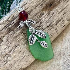 Pode incluir: Pingente artesanal com um peda&ccedil;o de vidro do mar em um tom verde vibrante. O pingente &eacute; adornado com um pingente de asas de anjo prateadas, uma conta facetada vermelha e um design de folha decorativa. O pingente pende de um cord&atilde;o fino e transparente.