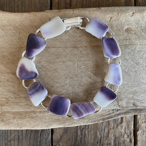 May include: A bracelet featuring ten smooth, irregularly shaped pieces of white and purple shell or stone, linked together by silver-toned metal rings and secured with a silver-toned metal box clasp closure, arranged in a circular formation.