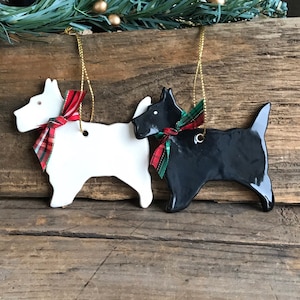 May include: Two ceramic dog ornaments, one white and one black, each with a red and green plaid ribbon bow. The ornaments are hanging from gold cords. The background is a rustic wooden surface, with greenery and gold ornaments.
