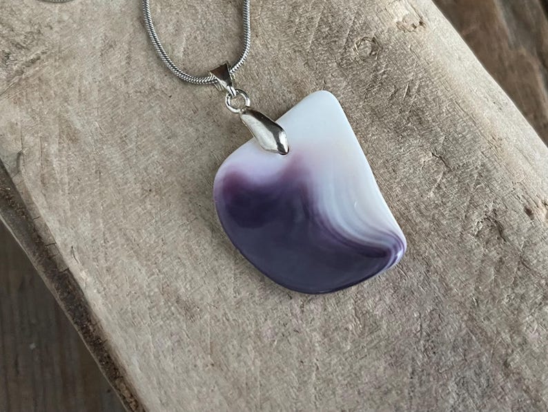May include: A pendant necklace featuring a polished, abstract-shaped stone with a gradient of white to deep purple hues. The stone is suspended from a silver-colored chain.