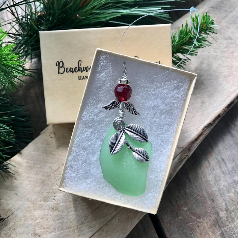 Pode incluir: Um enfeite artesanal com um pingente de vidro do mar verde claro, folhas de metal prateado e um motivo de anjo. Inclui uma conta vermelha e &eacute; apresentado em uma pequena caixa. A caixa tem o texto "Beachw" e "HAN".