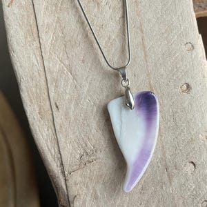 May include: A silver necklace with a pendant. The pendant is a heart-shaped piece, white at the base and transitioning to purple at the top. The necklace is displayed on a wooden surface.