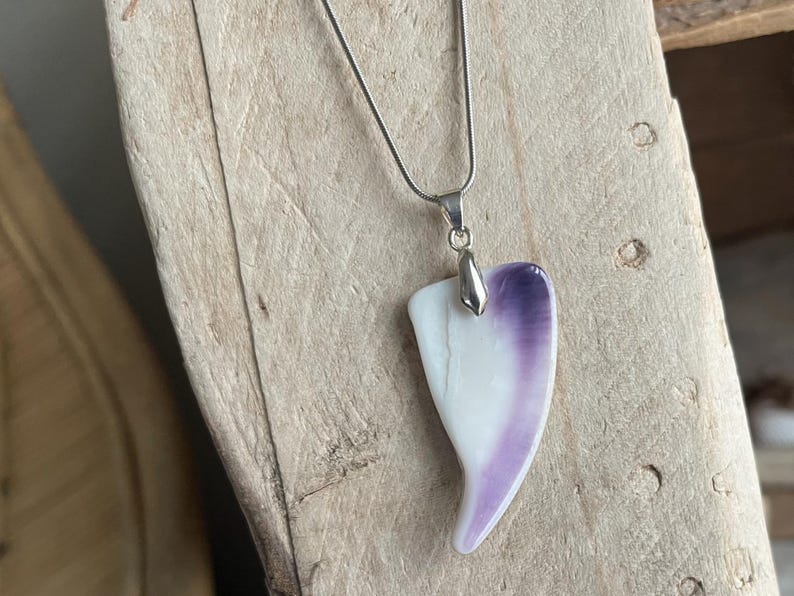 May include: A silver necklace with a pendant. The pendant is a heart-shaped piece, white at the base and transitioning to purple at the top. The necklace is displayed on a wooden surface.