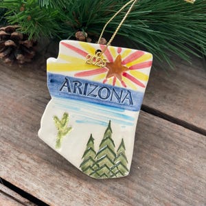 May include: A ceramic Arizona state ornament with a gold string for hanging. The ornament features a painted desert landscape with the word "ARIZONA" and the year "2025". The ornament is white with blue, yellow, red, and green accents.
