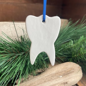 May include: A white, tooth-shaped ceramic ornament with a smooth, matte finish. The ornament features a small hole at the top, through which a bright blue ribbon is threaded for hanging. The design is simple, clean, and symmetrical.