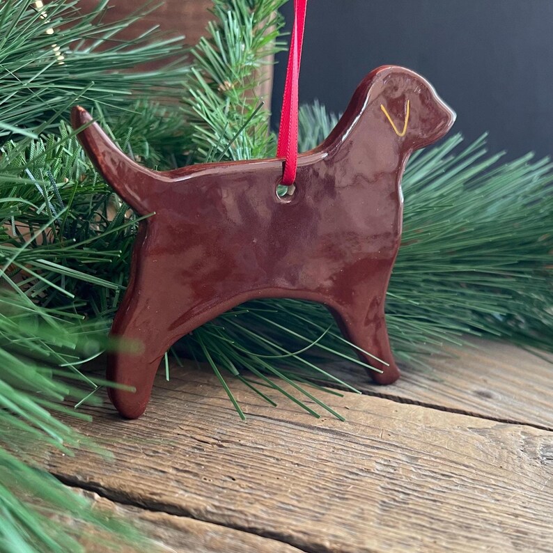 Pu&ograve; includere: Un ornamento in ceramica marrone scuro lucido a forma di cane labrador. La figura presenta una superficie liscia e riflettente con un piccolo foro sulla parte superiore per appenderla. Un sottile nastro rosso vivo &egrave; inserito nel foro.