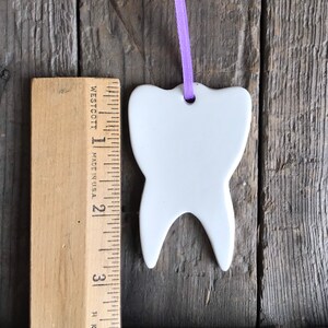 May include: A white ceramic tooth-shaped ornament with a purple ribbon loop for hanging, positioned next to a wooden ruler showing a height of approximately 3.5 inches from the top of the tooth to the bottom of the roots.