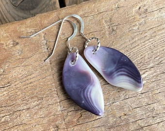 Pode incluir: Um par de brincos pendentes com pedras lisas em forma de gota, apresentando padrões de redemoinho em roxo escuro, lavanda e branco. As pedras estão suspensas em ganchos de metal prateado com um pequeno conector de anel torcido.