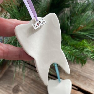 May include: A white ceramic tooth-shaped ornament with a smooth, glossy finish. A silver-toned metal charm displaying the year 2025 is attached to the top, suspended by a light purple ribbon. The ornament has two distinct roots at the base.