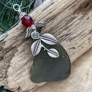 Pode incluir: Um pingente artesanal com um peda&ccedil;o de vidro do mar em tom verde escuro. O pingente &eacute; adornado com asas de anjo prateadas, uma conta vermelha e um design de folhas decorativas. O pingente pende de um cord&atilde;o transparente, sobre um fundo de madeira r&uacute;stica.