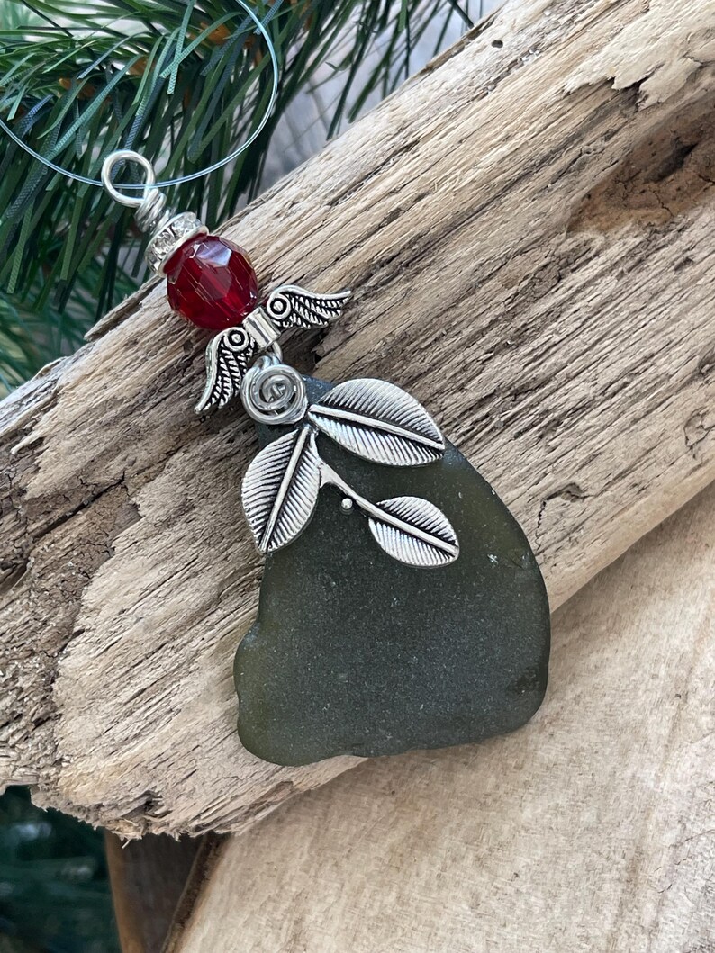 Pode incluir: Um pingente artesanal com um peda&ccedil;o de vidro do mar em tom verde escuro. O pingente &eacute; adornado com asas de anjo prateadas, uma conta vermelha e um design de folhas decorativas. O pingente pende de um cord&atilde;o transparente, sobre um fundo de madeira r&uacute;stica.