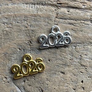 May include: Two metal charms shaped as the number 2026, one in a silver tone and one in a gold tone. Each charm features a small circular loop at the top for attachment, with the digits clearly formed in a continuous, raised metallic script style.