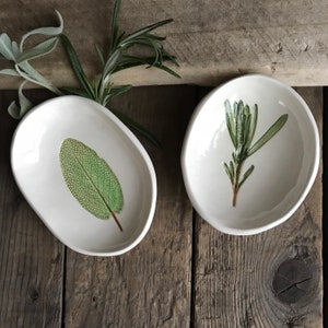 May include: Two white ceramic dishes with green leaf designs. The dish on the left has a single sage leaf design, while the dish on the right has a sprig of rosemary.