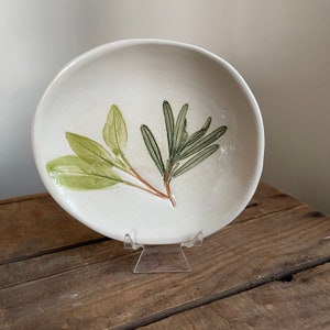 May include: A white ceramic plate with a hand-painted design of sage and rosemary sprigs. The sage leaves are light green, and the rosemary is dark green. The plate is displayed on a clear acrylic stand, set against a rustic wooden surface.