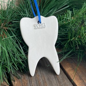 May include: A white ceramic tooth-shaped ornament with a smooth, glossy finish. A bright blue ribbon is threaded through a hole at the top, and a small silver-toned metal charm featuring the year 2026 hangs from the ribbon against the ornament.