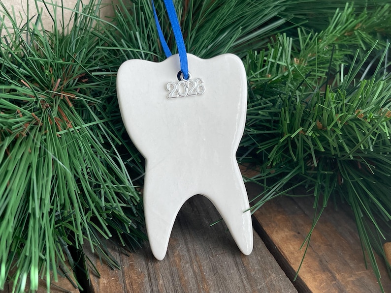 May include: A white ceramic tooth-shaped ornament with a smooth, glossy finish. A bright blue ribbon is threaded through a hole at the top, and a small silver-toned metal charm featuring the year 2026 hangs from the ribbon against the ornament.