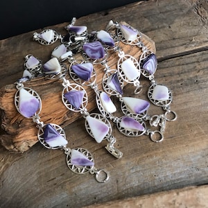 May include: Several silver-toned necklaces with oval and geometric links. Each link holds a polished stone in shades of white and purple. The necklaces are displayed on a wooden surface.