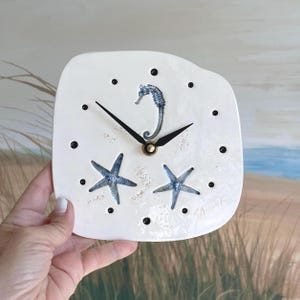 May include: A square-shaped white ceramic wall clock with rounded edges. The clock face features a blue seahorse and two blue starfish illustrations, accented with small black dots for hour markers and black clock hands with a gold center.