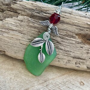 Pode incluir: Um pingente artesanal com um peda&ccedil;o de vidro do mar verde. O pingente &eacute; adornado com asas de anjo de metal prateado, uma conta facetada vermelha e detalhes de folhas. O pingente pende de um cord&atilde;o fino e transparente.