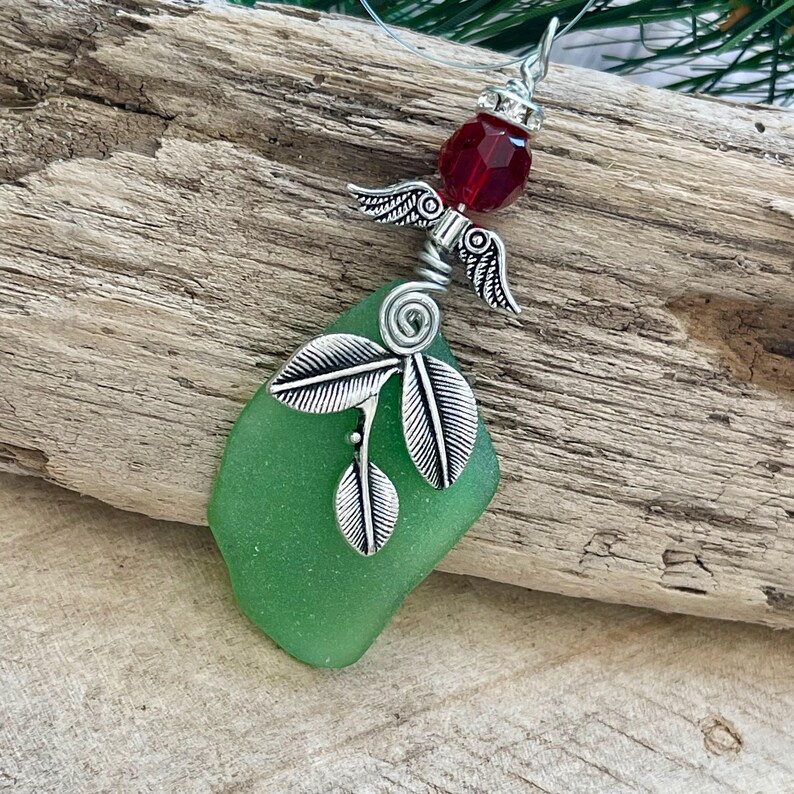 Pode incluir: Um pingente artesanal com um peda&ccedil;o de vidro do mar verde. O pingente &eacute; adornado com asas de anjo de metal prateado, uma conta facetada vermelha e detalhes de folhas. O pingente pende de um cord&atilde;o fino e transparente.