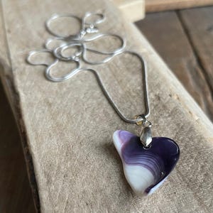 May include: A heart-shaped pendant necklace with a silver chain. The pendant features a swirl of white and purple colors. The necklace is displayed on a wooden surface.