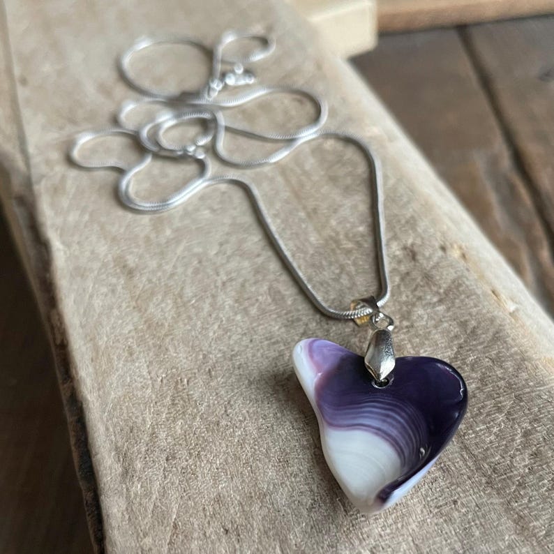 May include: A heart-shaped pendant necklace with a silver chain. The pendant features a swirl of white and purple colors. The necklace is displayed on a wooden surface.