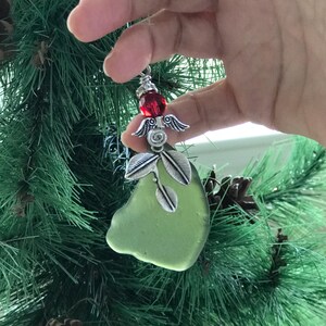 Pode incluir: Um enfeite de Natal artesanal com um peda&ccedil;o de vidro do mar verde claro. O enfeite &eacute; adornado com asas de anjo de metal prateado, folhas e uma vibrante conta vermelha. O enfeite &eacute; segurado em uma m&atilde;o, com uma &aacute;rvore de Natal verde ao fundo.