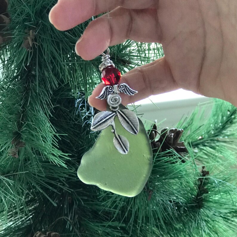 Pode incluir: Um enfeite de Natal artesanal com um peda&ccedil;o de vidro do mar verde claro. O enfeite &eacute; adornado com asas de anjo de metal prateado, folhas e uma vibrante conta vermelha. O enfeite &eacute; segurado em uma m&atilde;o, com uma &aacute;rvore de Natal verde ao fundo.