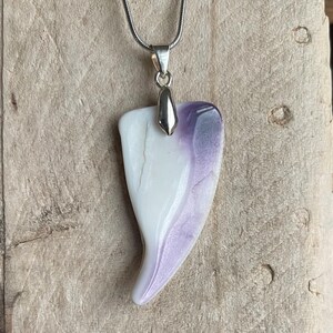 May include: A pendant necklace featuring a teardrop-shaped pendant with a white and purple marbled design. The pendant is suspended from a silver-colored chain. The necklace is displayed against a wooden background.