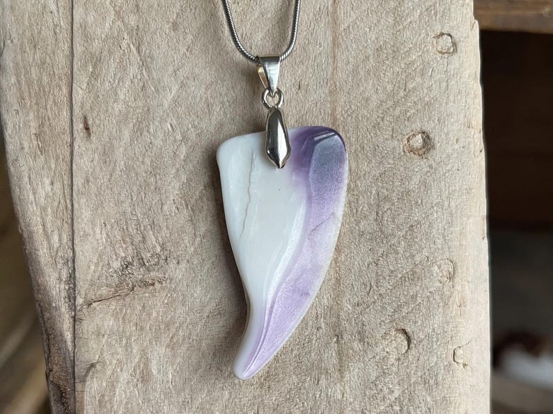 May include: A pendant necklace featuring a teardrop-shaped pendant with a white and purple marbled design. The pendant is suspended from a silver-colored chain. The necklace is displayed against a wooden background.