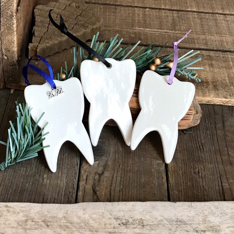 May include: Three white ceramic tooth-shaped ornaments with smooth, glossy surfaces. Each ornament hangs from a ribbon: one blue with a silver 2022 charm, one black, and one light purple. The ornaments are arranged in a row.