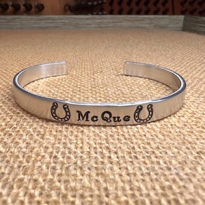 May include: A silver-toned metal cuff bracelet featuring an open design. The front surface is engraved with the text "Mc Que" centered between two horseshoe symbols. The bracelet has a smooth, polished finish and a curved, circular shape.