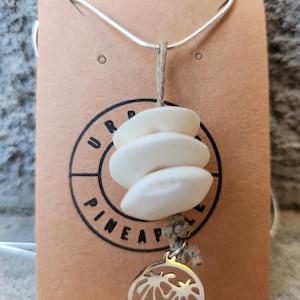 Puede incluir: Un collar con tres piedras blancas, lisas y redondeadas apiladas, suspendidas de un cordel de cáñamo beige, con un dije circular de metal plateado que representa dos palmeras, presentado sobre una tarjeta beige de Urban Pineapple.