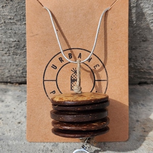 Puede incluir: Un collar de cadena de serpiente plateada con un colgante formado por cuatro discos circulares de cáscara de coco marrón oscuro apilados, suspendidos por un cordel de yute. Un pequeño dije circular de metal blanco con diseño de ola cuelga.