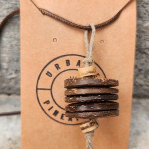 Puede incluir: Un collar con tres discos de madera de color marrón oscuro, circulares y texturizados, apilados verticalmente, acentuados por dos cuentas de madera más claras en un cordón de yute beige. El colgante se presenta en una tarjeta marrón con el texto URBAN PINE.