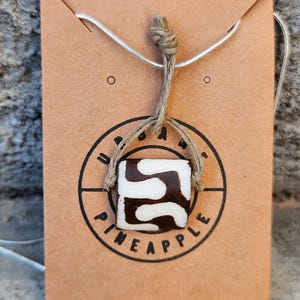 May include: A square-shaped pendant featuring a brown and white abstract pattern, suspended by a tan twine cord on a silver-colored chain. The pendant is displayed on a rectangular tan card printed with the text URBAN PINEAPPLE in a circle.