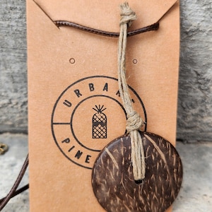 Puede incluir: Un colgante circular de cáscara de coco marrón oscuro suspendido de un cordón de yute color canela. El colgante está sujeto a una tarjeta de presentación rectangular de cartón marrón claro con un logo circular y el texto URBAN PINE.