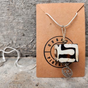 May include: A necklace featuring a silver-toned chain and a rectangular pendant with a black and white abstract pattern. A small circular charm with a wave design hangs from the bottom. The necklace is displayed on a brown card with the text URBAN PINEAPPLE.