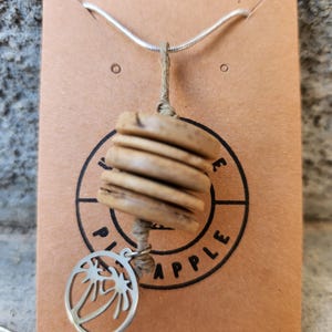 Può includere: Una collana con catenina argentata e un ciondolo composto da tre perline di legno circolari color marrone chiaro impilate su un cordino beige. Un piccolo ciondolo circolare argentato con il disegno ritagliato di due palme pende in basso.