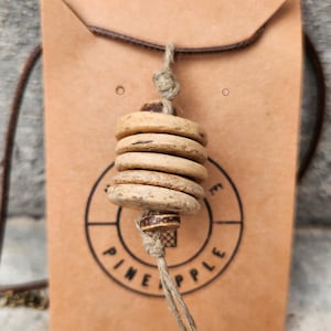 Op de afbeelding: Een ketting met een stapel van vier lichtbruine, ronde, getextureerde kralen geregen aan een natuurlijk touwtje. Het touwtje is boven en onder de kralen geknoopt en het geheel wordt gepresenteerd op een beige kaart met de tekst PINEAPPLE.