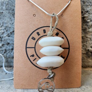 Peut inclure: Un collier composé de trois pierres blanches, lisses et ovales empilées, suspendues à un cordon en ficelle beige. Une petite breloque circulaire en métal argenté avec un motif de vague est fixée en bas. Le collier est présenté sur une carte brune avec le texte URBANE PIECE.